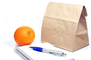 Brown paper lunch bag and orange next to notebook & pen
