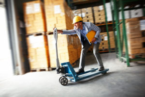 Logistics, Hard-hatted worker happily riding pallet jack in warehouse towards loading dock