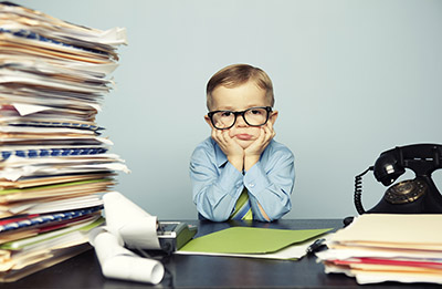 Paperwork Stress, Young boy in large glasses dressed as if adult, sitting head in hands at desk full of paperwork