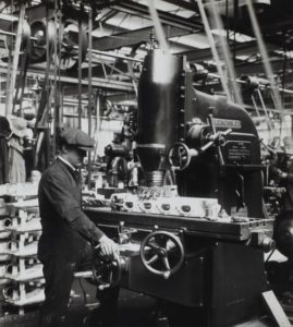 Manufacturing History: Old photo of male factory worker in hat and jacket at machine.