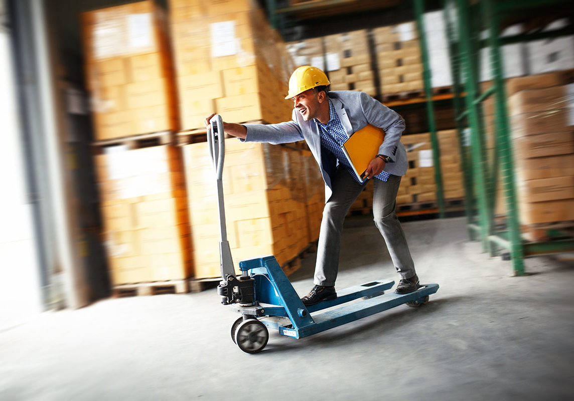 Logistics, Hard-hatted worker happily riding pallet jack in warehouse towards loading dock